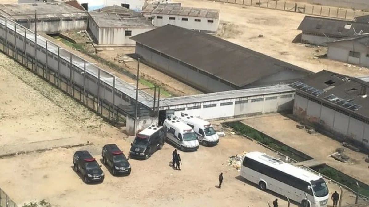 Portão de unidade prisional com agentes penitenciários ao fundo