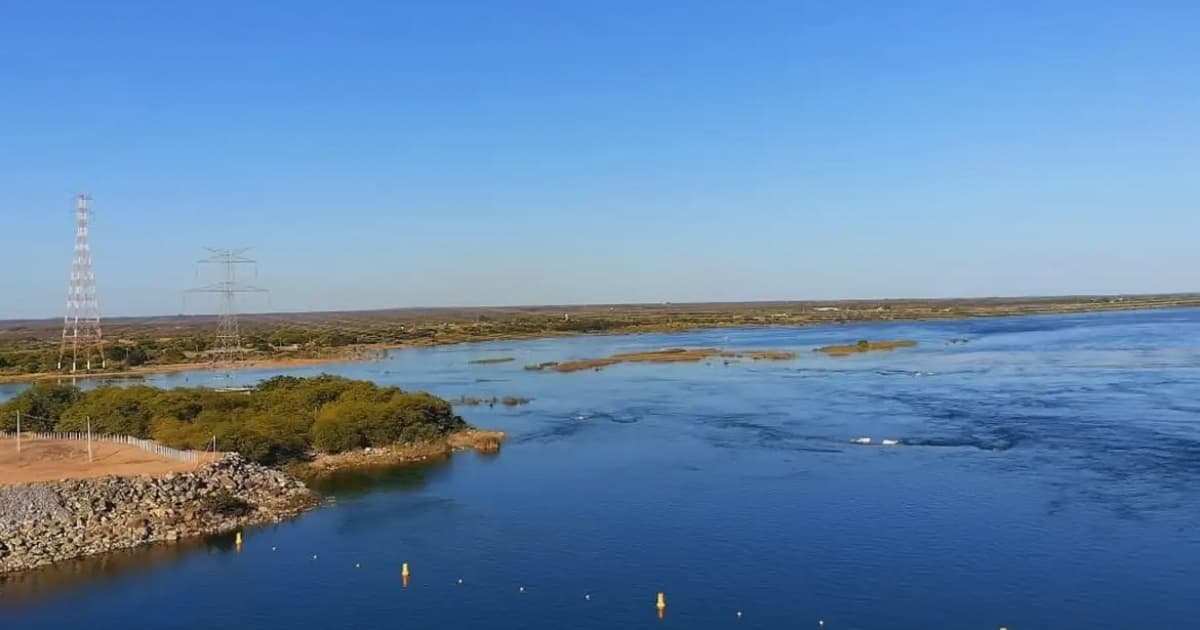 Barragem com estrutura de concreto vista de frente, represando volume de água em área rural